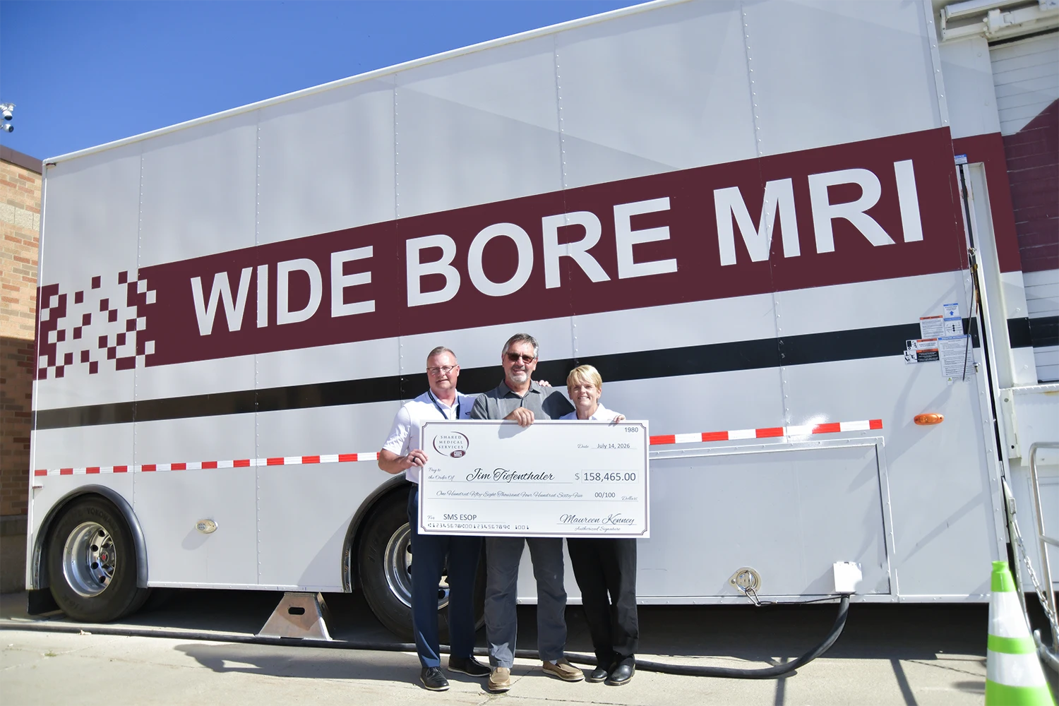 Shared Medical Services colleagues present retired Quick Responder Driver Jim Tiefenthaler with a large ceremonial ESOP check in front of a mobile MRI scanner.